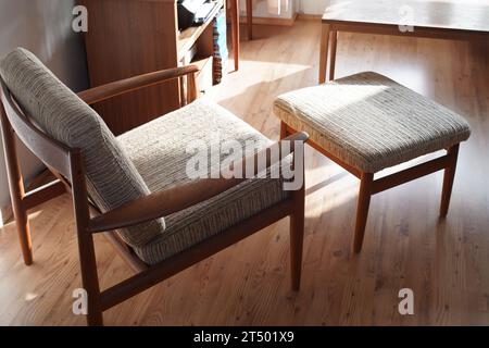 Ein Mid Century Sessel mit Ottoman aus Teakholz im dänischen Design in einem gemütlichen, modern eingerichteten Wohnzimmer im Stil der 60er Jahre mit weißer Wand und Fernseher Stockfoto