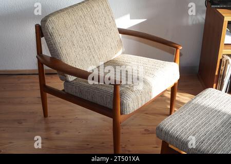 Ein Mid Century Sessel mit Ottoman aus Teakholz im dänischen Design in einem gemütlichen, modern eingerichteten Wohnzimmer im Stil der 60er Jahre mit weißer Wand und Fernseher Stockfoto