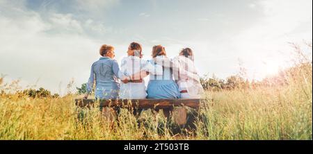 Vier lächelnde Frauen kleideten leichte Sommerkleidung, wenn sie auf der Wiesenbank sitzen und draußen spazieren gehen. Freundschaft der Frau, Beziehungen und Stockfoto