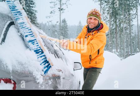 Ein lächelnder Mann in einem lustigen nepalesischen Hut, der Schnee mit einer großen Schneebürste aus dem Auto entfernt. Das Auto friert auf der Landstraße des Waldhauses. Gewinnen Stockfoto