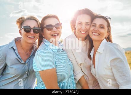 Porträts in halber Länge von vier fröhlichen, lächelnden Frauen in Sonnenbrille, die sich umarmen und beim Wandern im Freien in der hellen Sonne in die Kamera schauen. frauenfreitag Stockfoto