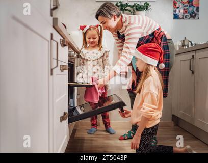 Süße kleine Mädchen in roten Weihnachtsmannmützen mit Mutter, die selbstgemachte süße Kekse macht. Sie legen Backpapier in den Ofen. Vorbereitung von Weihnachtskeksen. Stockfoto