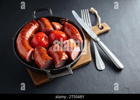 In Scheiben geschnittene frittierte Schweinefleischwurst und gegrillte Tomaten auf einer schwarzen gusseisernen Pfanne auf einem Holzbrett auf einem schwarzen Steintisch Stockfoto