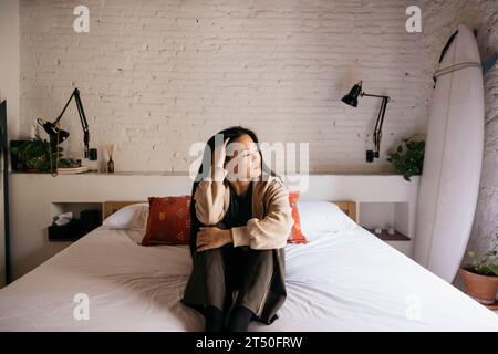 Nachdenkliche junge, schöne Frau, die auf dem Bett sitzt Stockfoto