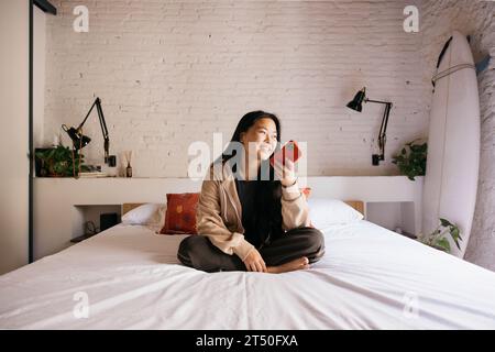 Lächelnde junge, schöne Frau, die auf dem Bett sitzt und eine Sprachnachricht mit einem roten Telefon sendet Stockfoto