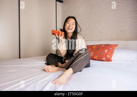 Lächelnde junge, schöne Frau, die auf dem Bett sitzt und eine Sprachnachricht mit einem roten Telefon sendet Stockfoto