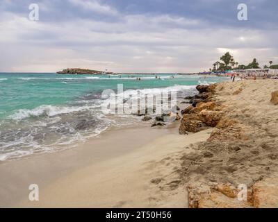 Menschen, die an bewölkten Tagen in Meereswellen schwimmen. Malerischer Nissi-Strand mit turbulentem Cyan-Wasser, eine kleine felsige Insel, bewölkter Himmel in Ayia Napa, Zypern Stockfoto