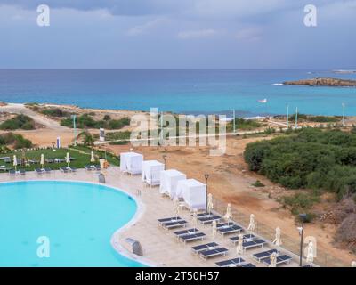 Blick von oben auf einen leeren Swimmingpool mit Sonnenliegen und Sonnenschirmen, das Mittelmeer mit einer kleinen Insel in der Nähe des Nissi Strandes und die Wanderpromenade entlang der Küste Stockfoto