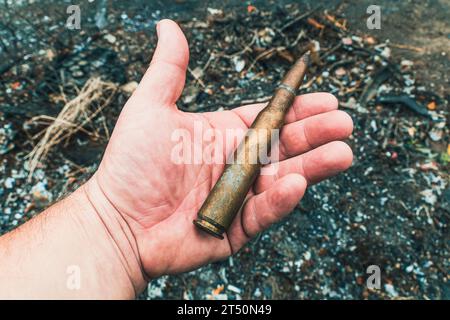 Eine großkalibrige Maschinengewehrpatrone in der Hand vor dem Hintergrund verbrannter Erde. Nahaufnahme Stockfoto