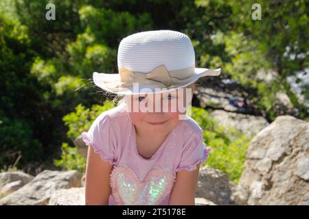 Ein fünfjähriges Mädchen mit weißem Hut lächelt. Sie trägt ein rosafarbenes T-Shirt, grüne Bäume im Hintergrund, blaue Augen, blondes Haar, Stockfoto