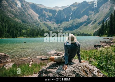 Der Mann sitzt und bewundert den Alpensee vor ihm mit mehreren Wasserfällen in der Ferne Stockfoto