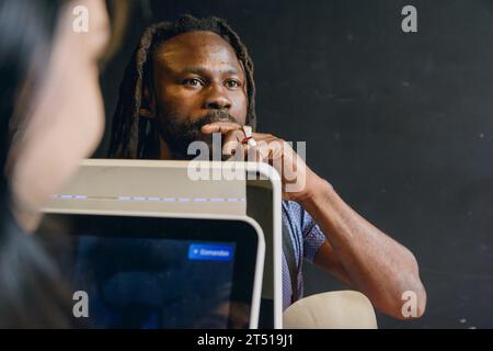 Junger Mann afrikanischer Ethnie mit Dreadlocks und Bart im Restaurant, der vor dem Check-out-Computer steht, mit der Hand auf dem Gesicht wie ein Stockfoto