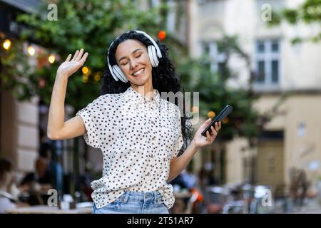 Junge schöne hispanische Frau hört Musik, singt und tanzt, während sie in der abendlichen Stadt spaziert, Frau mit lockigem Haar benutzt Kopfhörer und App auf dem Telefon. Stockfoto