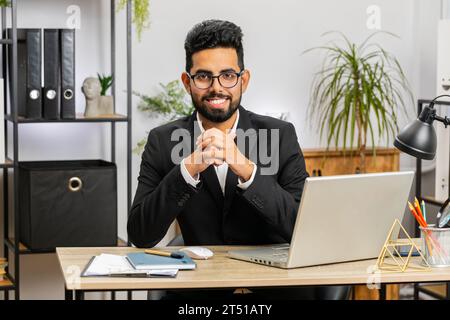 Porträt eines glücklichen lächelnden bärtigen indischen Geschäftsmannes am modernen Schreibtisch des Heimbüros, der in die Kamera schaut. Männlicher arabischer Hindu in formellem Anzug. Schöner Mann, der im Internet an einem Laptop arbeitet Stockfoto