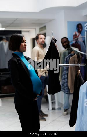 Schwangere asiatische Kundin untersucht Kleiderständer und hält Jacke am Kleiderbügel im Kaufhaus. Zukünftige Mutter, die den Bauch hält und Mutterschaftskleidung in der Modeboutique wählt Stockfoto