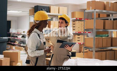 Zwei Frauen mit Schutzhelden organisieren Produkte in Regalen, schauen sich Tablets an, um die Lagerlogistik zu überprüfen und den Vertrieb zu planen. Lagerleiter und Mitarbeiter zählen Papppackungen. Stockfoto