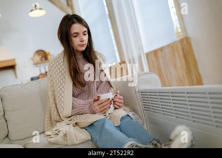 Junge Frau mit Karo in der Nähe der elektrischen Heizung, die eine Tasse heißes Getränk hält, während sie zu Hause auf dem Sofa sitzt. Stockfoto