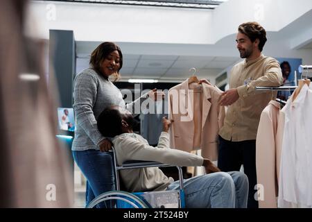 Mann mit körperlicher Behinderung, der Jacke mit Freundin beim Einkaufen im Bekleidungsgeschäft wählt. Kaufhaus afroamerikanischer Kunde im Rollstuhl, der ein Outfit mit Frau auswählt Stockfoto