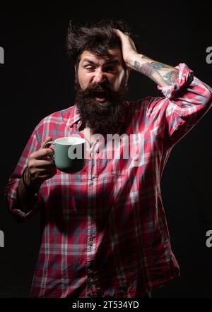 Mans hält eine Tasse Kaffee und Gähnen auf Schwarz. Morgentee. Gähngesicht. Mann mit Teetasse. Hipster-Mann gähnt mit einer Tasse Kaffee. Bärtiger Mann Stockfoto