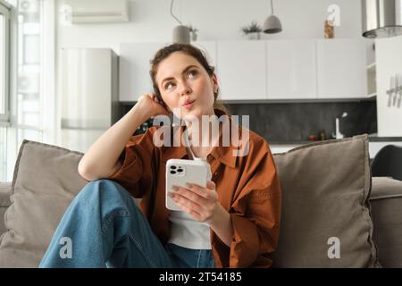 Porträt einer niedlichen neugierigen Frau, die mit dem Smartphone auf dem Sofa sitzt, aufblickt und nachdenkt, eine Bestellung aufgibt, Online-Shopping über die Handy-App Stockfoto