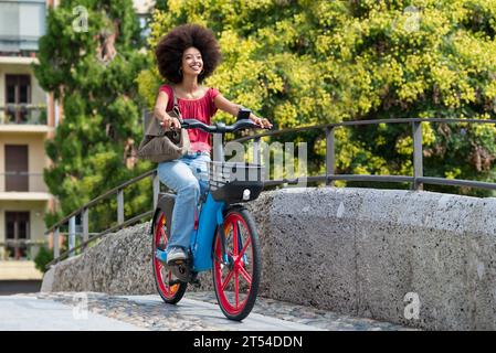 Seitenansicht einer lächelnden jungen marokkanischen Frau mit Handtasche, die wegschaut, während sie mit dem Elektrofahrrad auf der Stadtbrücke mit Geländer gegen verschwommenes Grün fährt Stockfoto