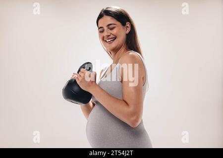 Schwangere Frau im dritten Trimenon hebt eine Kettlebell in einem gut ausgestatteten Fitnessstudio. Sie legt Wert auf ihr Wohlbefinden und nähert sich der Mutterschaft Stockfoto