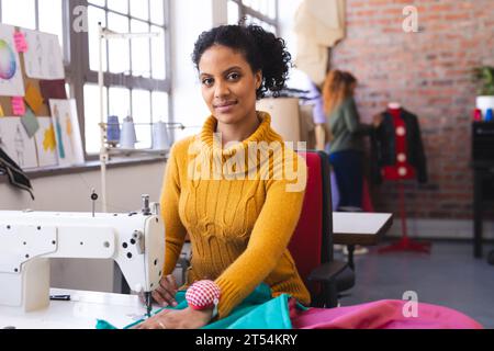 Glückliche birassische Modedesignerin mit Nähmaschine im sonnigen Studio Stockfoto