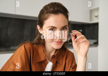 Porträt einer schönen jungen Frau, lächelnd und glücklich aussehend, zieht ihre Brille ab und blickt in die Kamera, sitzt zu Hause in der Küche Stockfoto