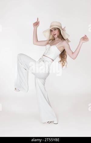 Eine verspielte junge Frau, die auf einem Bein steht, die Arme hochgehoben hat und die Kamera auf weißem Hintergrund betrachtet. Blonde Frau in voller Länge, die einen Floppy Strohhut trägt Stockfoto