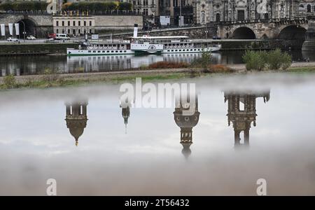 03. November 2023, Sachsen, Dresden: Die Türme des Ständehauses (l-r), des Georgentors, des Hausmannsturms und der Hofkirche spiegeln sich in einer Pfütze an der Königsufer gegenüber der Altstadt an der Elbe, der historische Raddampfer der Sächsischen Dampfschiffsgesellschaft hat am Terrassenufer angedockt. Wind und Regen dominieren das Wetter in Deutschland am Wochenende. Vereinzelte Gewitter an der Nordsee sind nicht auszuschließen, wie der Deutsche Wetterdienst am Freitag in Offenbach berichtete. Auch der Einfluss des Niederdrucks wird in der kommenden Woche nicht nachlassen: Er wird teilweise mild, nass und stürmisch bleiben. Stockfoto