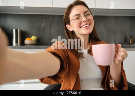 Sorgloses Mädchen in Brille, nimmt Selfie mit Smartphone, lächelt und sieht glücklich aus, genießt heißes Getränk in der Küche zu Hause Stockfoto