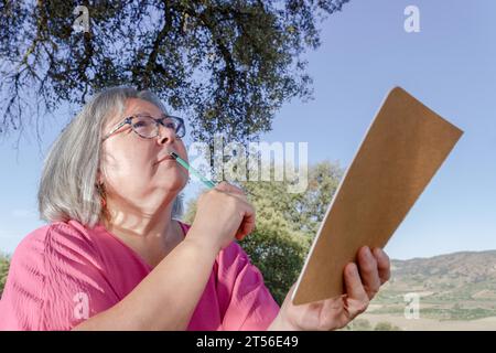 Ältere weißhaarige Frau mit einem Bleistift auf den Lippen und einem Notizbuch in der Hand, nachdenklich und träumend, blickend in den Himmel auf dem Feld Stockfoto