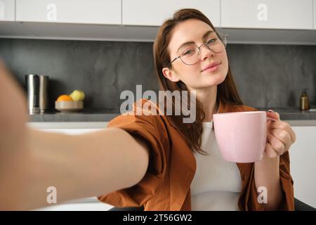 Porträt einer schönen jungen Frau in Brille, macht Selfie mit einer Tasse Tee, sitzt in der Küche, hält Smartphone Stockfoto