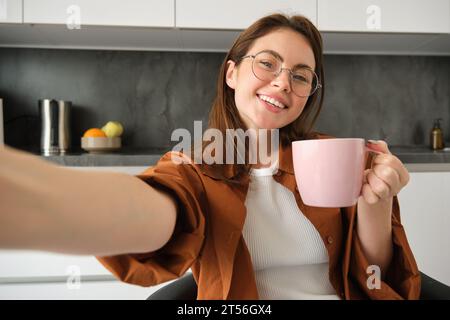 Porträt einer glücklichen jungen Frau in Gläsern, nimmt Selfie mit Smartphone, trinkt Kaffee, posiert mit einer köstlichen Tasse Tee in der Küche Stockfoto