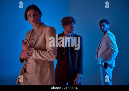 Drei junge, elegante Männer posieren auf blauem Hintergrund in eleganten Anzügen, blauen Lichtern, Modekonzept Stockfoto