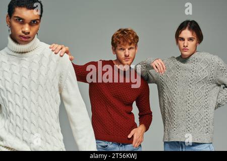 Gut aussehende, vielfältige Männer in Pullover mit Händen auf den Schultern und Blick auf Kamera, Mode Stockfoto