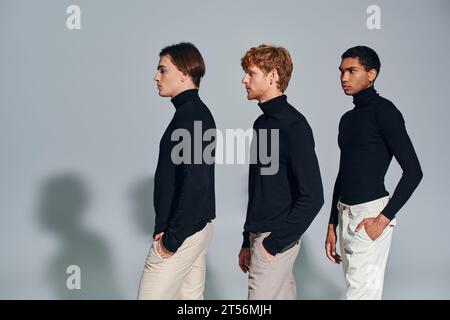 Drei multiethnische Männer mit schwarzen Rollkragenbündchen, die in einer Akte vor grauem Hintergrund laufen, Modekonzept Stockfoto