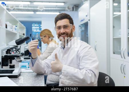 Zufrieden mit den Forschungsergebnissen zeigt der Laborassistent der Wissenschaftler die Daumen nach oben zur Kamera, ein erfahrener Labormitarbeiter verwendet das Mikroskop im Gebäude Stockfoto