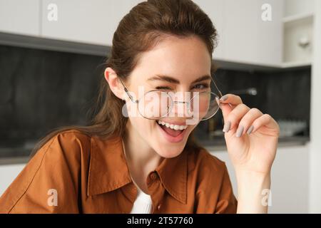 Porträt einer schönen jungen Frau, lächelnd und glücklich aussehend, zieht ihre Brille ab und blickt in die Kamera, sitzt zu Hause in der Küche Stockfoto