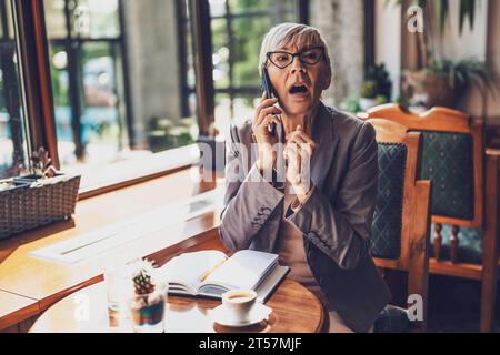 Reife Frau sitzt im Café und entspannt sich. Sie trinkt Kaffee und spricht am Telefon. Stockfoto