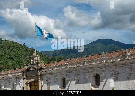Guatemala, La Antigua - 20. Juli 2023: Kolonialkunstmuseum Teil der Universität: Nordwand mit Nationalflagge. Oben, Landschaft mit Dschungel bedeckt Vol Stockfoto