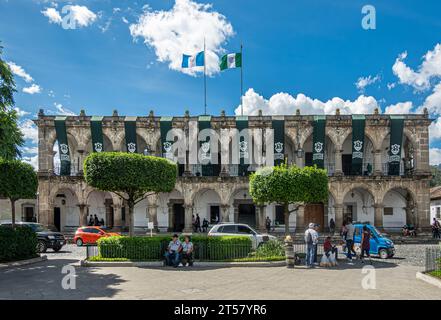 Guatemala, La Antigua - 20. Juli 2023: Historisches Rathaus mit Nationalflagge und Stadtflagge, die vor blauer Wolkenlandschaft winkt. Autos und Leute auf plaz Stockfoto