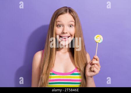 Porträt des erstaunten überfröhlichen Teenager-Mädchens mit langen Haarfrisuren tragen bunter Singlet halten Lutscher in der Hand isoliert auf lila Hintergrund Stockfoto