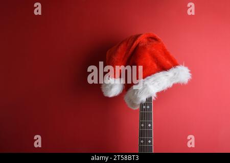 Akustikgitarrenhals mit Weihnachtsmann-Hut auf dem Kopf, der an einer isolierten roten Wand gelehnt ist. Weihnachtsgitarrenmusik-Konzept. Stockfoto