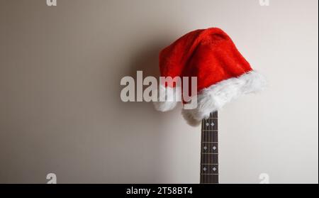 Akustikgitarrenhals mit Weihnachtsmann-Hut auf dem Kopf, der an einer isolierten weißen Wand gelehnt ist. Weihnachtsgitarrenmusik-Konzept. Stockfoto