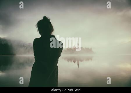 Frau im grauen Mantel, die einen Sonnenaufgang im Nebel am Ufer des Bleder Sees beobachtet, Slowenien III Stockfoto