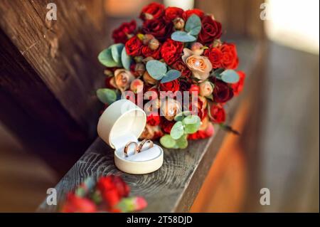Eheringe in der Nähe von rotem Blumenstrauß mit Rosen auf hölzernem Hintergrund. Brautaccessoires. Das Konzept einer Hochzeit im ethnischen Stil Stockfoto