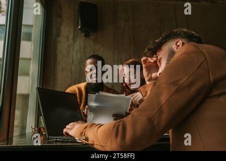 Müder Geschäftsmann, der auf dem Schoß arbeitet, während seine multirassischen Kollegen Papierkram lesen Stockfoto