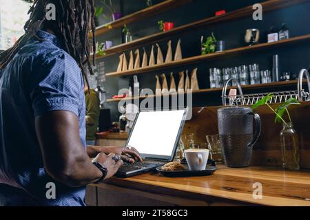 Rückansicht eines hübschen schwarzen Geschäftsmannes mit Dreadlocks, der freiberuflich an seinem Laptop im Restaurant arbeitet und sein Geschäft online verwaltet, während h Stockfoto