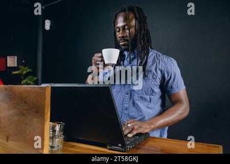 Schwarzer Mann afrikanischer Ethnie, mit Dreadlocks und lässiger Kleidung, stehend und mit seinem Laptop arbeitend, leitete sein Geschäft, im Restaurant und im Hafen Stockfoto